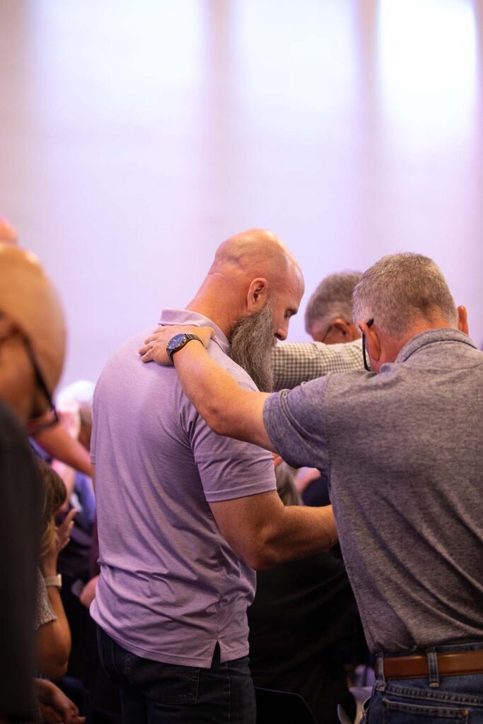 bald, bearded man, praying with another man in a room of small groups praying together